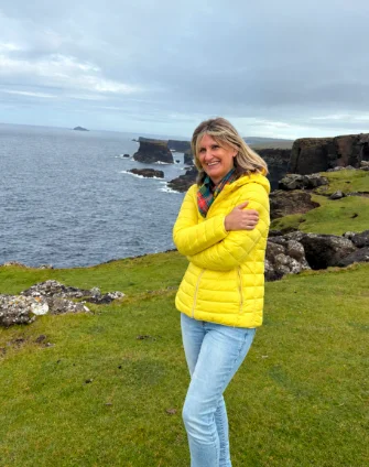 Susanne Eichhorn in gelber Jacke steht lächelnd auf einer grünen Klippe am Meer